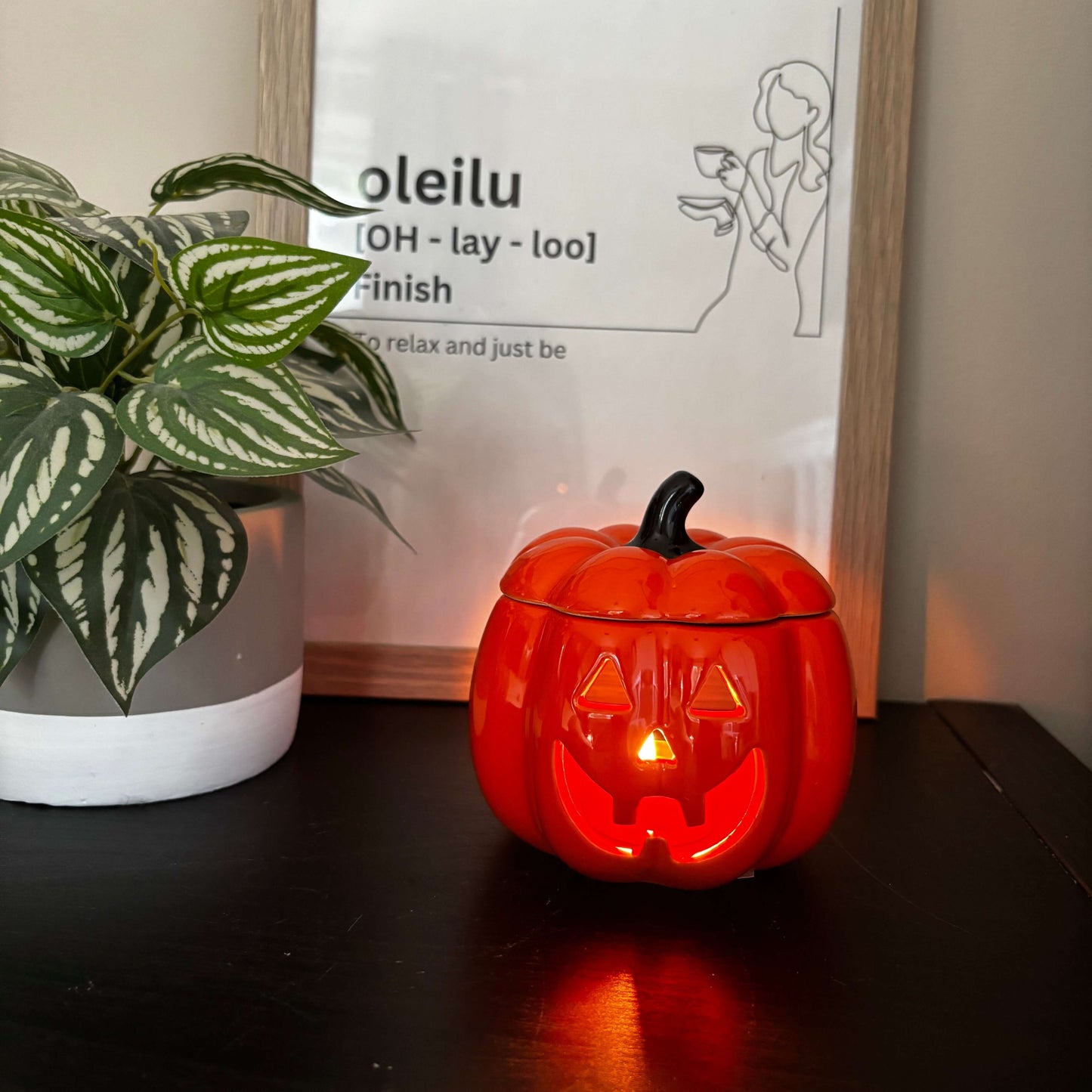 Small pumpkin-shaped lantern with a lit candle inside on a surface next to a framed picture and plant.