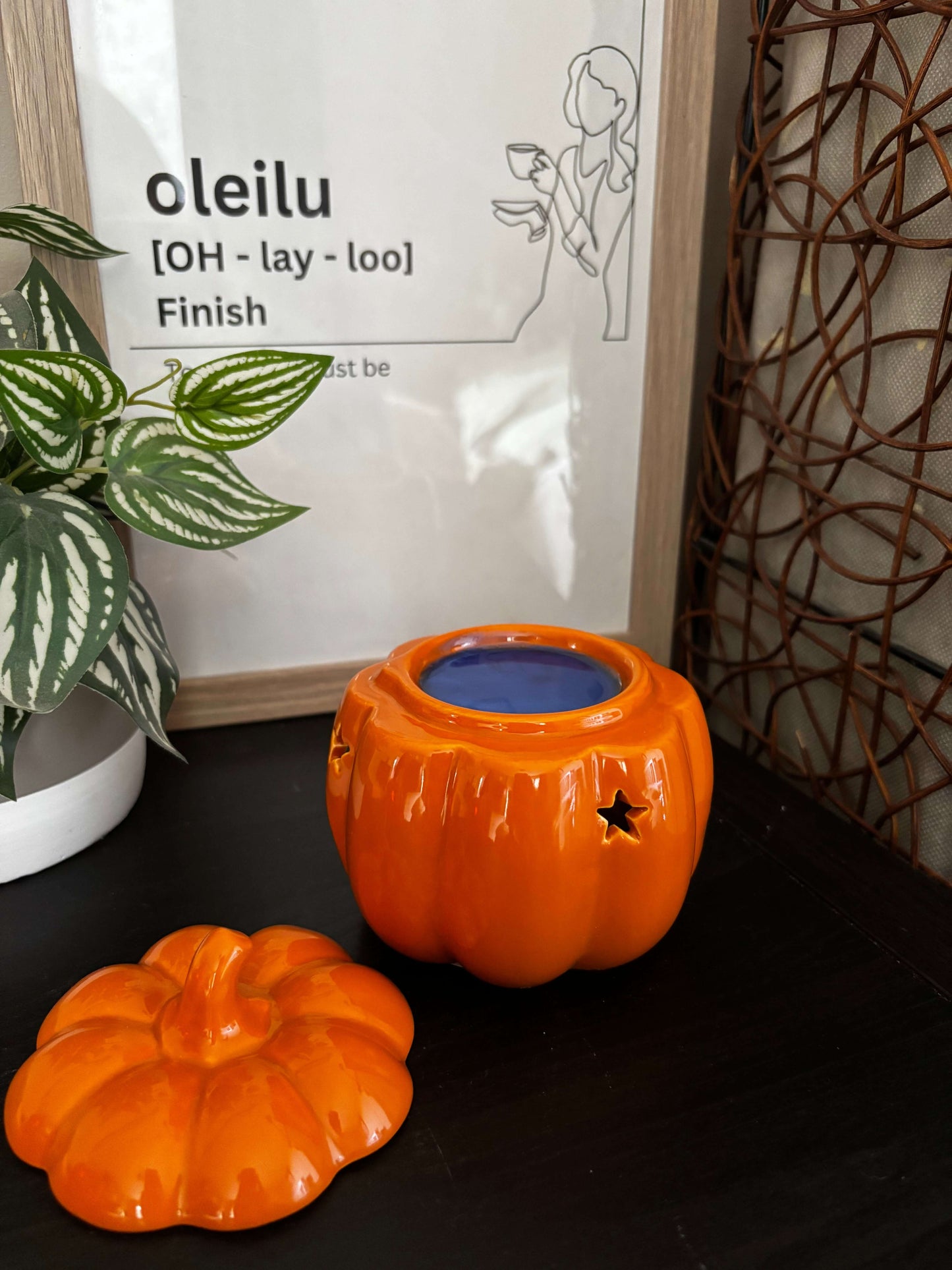 Two orange pumpkin-shaped candles on a surface with a plant and a product packaging in the background.