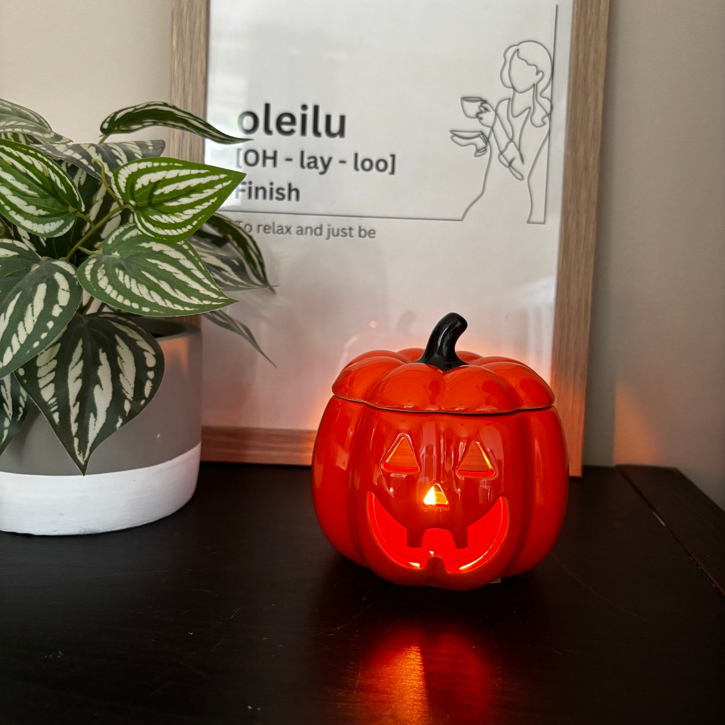 Small pumpkin-shaped lantern with a lit candle inside on a surface next to a framed picture and plant.