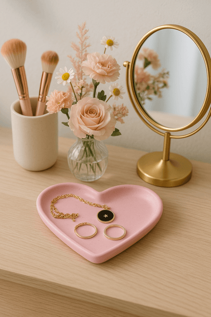 Heart-shaped jewelry tray with rings, gold necklace, makeup brushes, and flowers on a wooden surface.