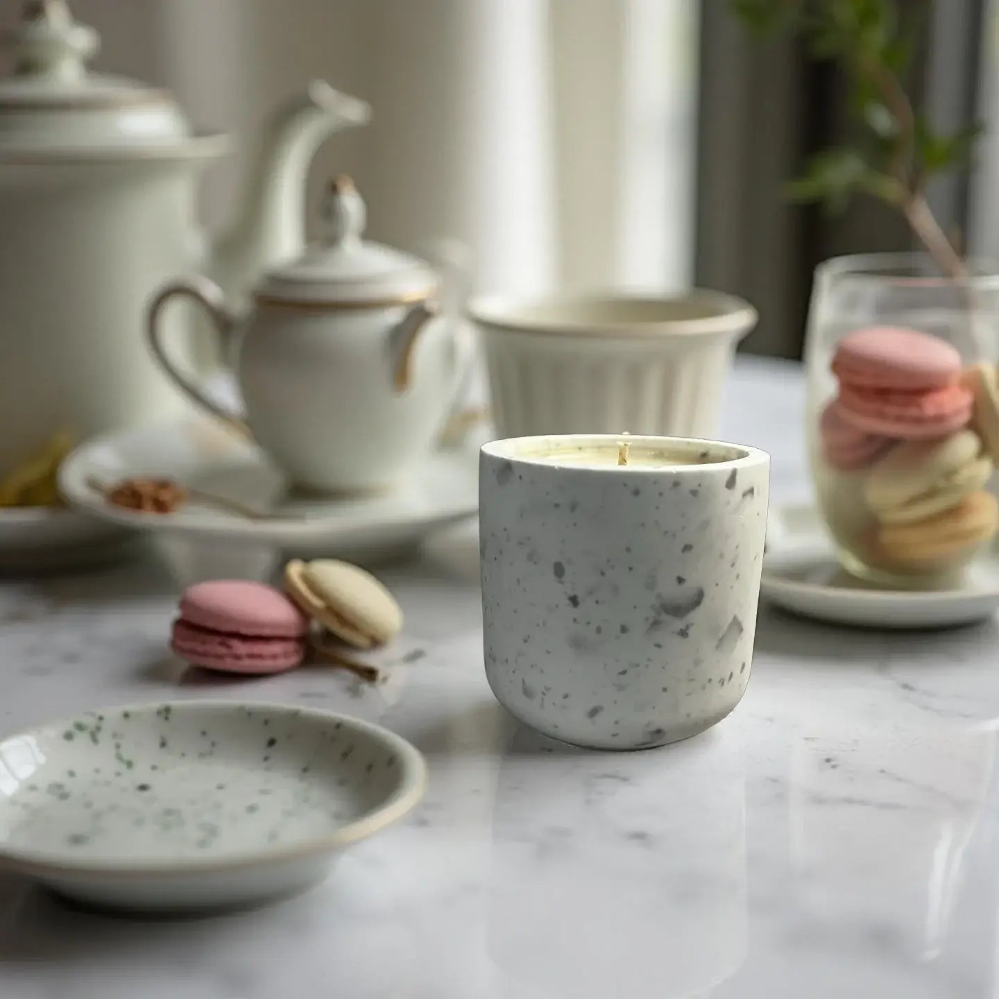Handmade Afternoon Tea candle surrounded by delicate macarons and elegant tea set on a marble table.